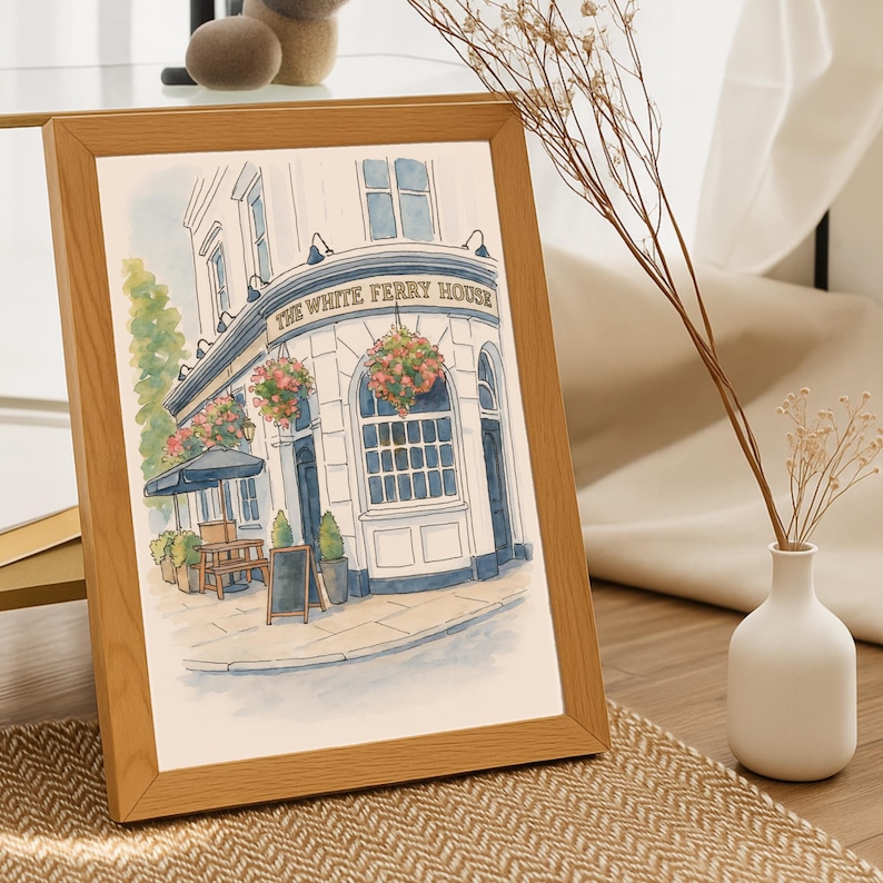 May include: A framed watercolor print of a pub, "The White Ferry House", with a light brown wooden frame. The building is white with blue accents, floral arrangements, and a black awning. A small chalkboard stands in front.