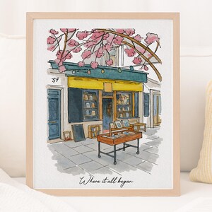 May include: Watercolour print of a bookshop with a yellow awning and blue doors, framed in light wood. Cherry blossoms bloom overhead. The text reads "Where it all began."