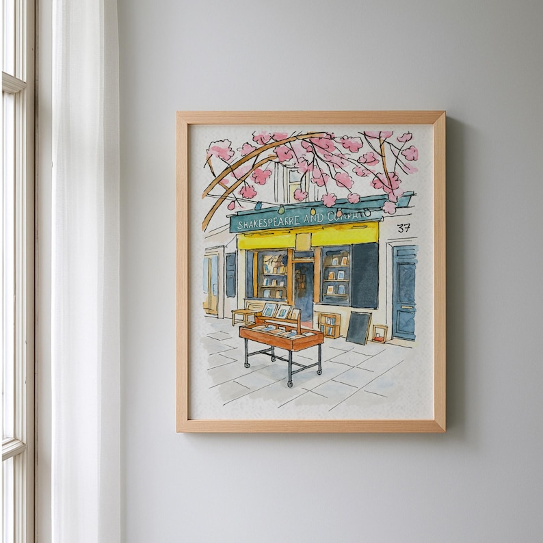 May include: Watercolor illustration of a bookstore, framed in light wood. The shop has a yellow awning with the text "Shakespeare and Co." and a display of books outside. Pink blossoms arch over the building.