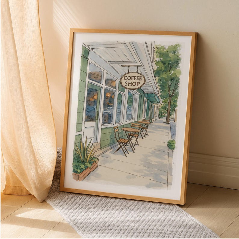 May include: Watercolor print of a coffee shop with a green facade, tables, and chairs on the sidewalk. The sign reads "Coffee Shop". The artwork is framed in a light brown wooden frame.