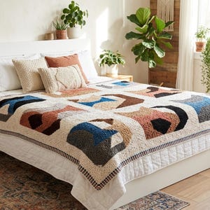 Może przedstawiać: Ręcznie robiona kołdra z geometrycznym wzorem w odcieniach kremu, brązu, błękitu i czerni, wyeksponowana na łóżku z pasującymi poduszkami. Kołdra ma powtarzający się wzór i jest obramowana ciemnoniebieskim i białym wykończeniem. Łóżko znajduje się w dobrze oświetlonym pokoju.