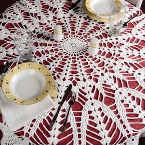 May include: A white, crocheted lace tablecloth covers a round table, with a red underlay visible through the openwork design. The table is set with plates, silverware, glasses, and salt and pepper shakers, creating a dining setting.