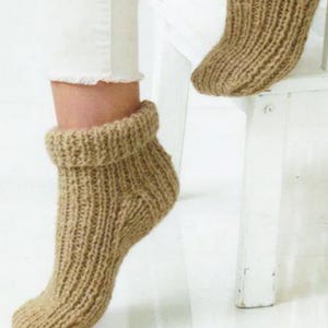 May include: Close-up of a pair of tan knitted socks with a ribbed design. The socks have a folded-over cuff and are displayed on a white surface. The texture of the yarn is visible.