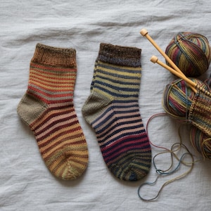Peut inclure: Deux chaussettes tricotées à la main avec des rayures horizontales dans les tons de marron, rouge, jaune et bleu. À côté des chaussettes se trouvent des pelotes de laine aux couleurs assorties, des aiguilles à tricoter et un petit morceau de tissu tricoté.