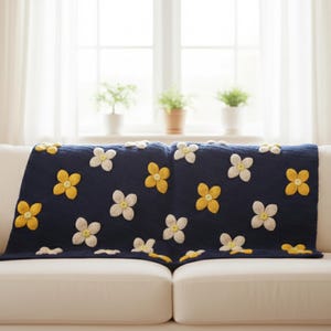 May include: A navy blue throw blanket with a floral pattern of yellow and white flowers, draped over a white sofa. The blanket is made of a soft material and adds a touch of color to the room. The background includes a window with white curtains and potted plants.