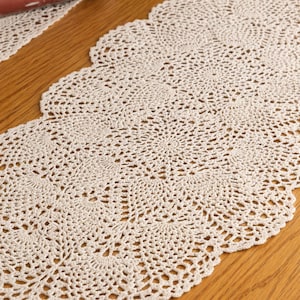 May include: Cream-colored, crocheted table runner with a scalloped edge. The runner is made of a delicate, open-weave design, showcasing intricate patterns. The runner is placed on a wooden surface.
