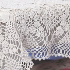 Könnte beinhalten: Nahaufnahme einer cremefarbenen, gehäkelten Tischdecke. Das komplizierte Spitzendesign weist florale und geometrische Muster auf. Die Ränder der Tischdecke sind gewellt, was einen zarten Touch verleiht. Die Textur des Häkels ist sichtbar und unterstreicht die Handwerkskunst.