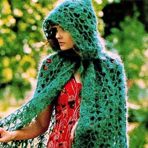 May include: A green, open-weave crochet hooded cape with fringe. The cape is worn over a red floral dress. The crochet work has a textured, open design. The background is a blurred view of green foliage.