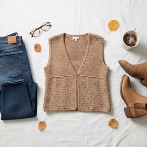 Op de afbeelding: Een herfst-thema flat lay met een beige gehaakt vest, donkerblauwe jeans, bruine enkellaarsjes en een kop koffie. De scène bevat een bril en gevallen bladeren, wat een gezellige, seizoensgebonden uitstraling creëert.