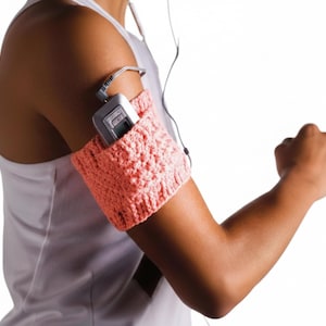 May include: A coral-colored, knitted armband holds a silver MP3 player. The armband is worn on the upper arm, with the player's cord and earbuds visible. The person is wearing a white tank top.