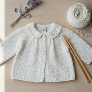 May include: A cream-coloured, knitted baby cardigan with a scalloped collar and button closure. The garment is displayed with a ball of white yarn, knitting needles, and a bundle of lavender tied with a blue ribbon. The cardigan has a delicate, openwork pattern.