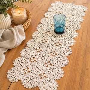 May include: A cream-colored, open-weave lace table runner with a floral pattern, placed on a wooden table. A blue glass and a candle in a woven holder are also on the table, along with a white vase and a beige cloth.
