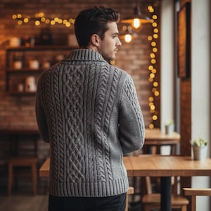 Op de afbeelding: Een grijze kabelgebreide trui met een sjaalkraag. De trui heeft een textuurpatroon en wordt gedragen door een persoon in een café. De achtergrond omvat houten tafels, stoelen en warme verlichting.