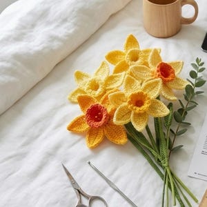 May include: A bouquet of handmade crochet daffodils in shades of yellow and orange, with green stems and leaves. The flowers are arranged on a white surface, alongside a wooden mug, a tablet, and crafting tools.
