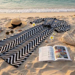May include: A black and white chevron patterned maxi dress, straw hat with black ribbon, sunglasses, woven tote bag, and open book on a sandy beach. The scene includes seashells and flowers.