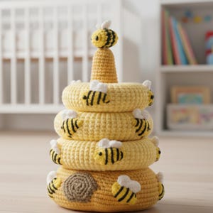 Könnte beinhalten: Ein handgefertigtes, gelbes Häkel-Stapelspielzeug in Form eines Bienenstocks. Das Spielzeug besteht aus einer Reihe gelber Ringe mit Applikationen von schwarzen und gelben Bienen. Eine kleine Biene sitzt auf der kegelförmigen Mitte. Die Basis hat ein braunes Wabenmuster.