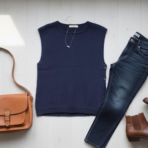 May include: A navy blue sleeveless sweater, dark wash jeans, and brown leather ankle boots are arranged on a light wood surface. A brown leather satchel bag and a silver necklace complete the outfit. The items are arranged to suggest a casual, everyday style.
