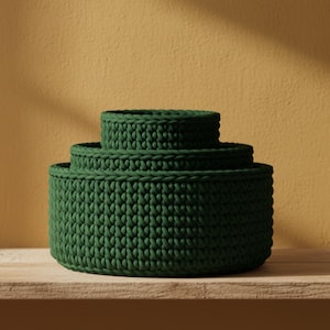 Puede incluir: Tres cestas redondas de ganchillo verde oscuro de diferentes tamaños apiladas sobre una estantería de madera. Las cestas tienen un aspecto texturizado y probablemente se utilizan para guardar o decorar. El fondo es amarillo cálido.