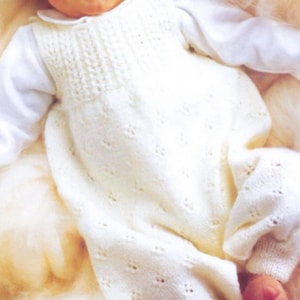 May include: A white knitted baby romper with a textured bodice and delicate openwork detailing. The romper is paired with a white long-sleeved shirt. The outfit is designed for a baby and is made of soft, comfortable material.