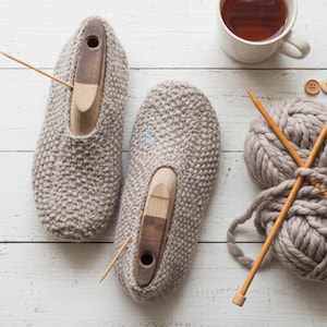 May include: A pair of grey knitted slippers with wooden shoe lasts, a ball of grey yarn, knitting needles, and a cup of tea. The items are arranged on a white wooden surface, evoking a handmade, cosy atmosphere.