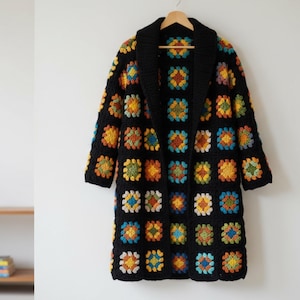 May include: A long, black crochet coat with a shawl collar. The coat features colorful square patterns in shades of yellow, orange, blue, and green. The coat is hanging on a wooden hanger.
