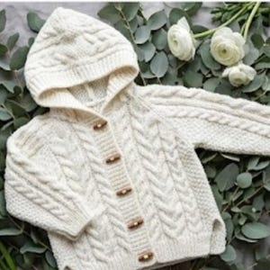 Może przedstawiać: Kremowy sweter z kapturem dla dziecka z warkoczowym wzorem. Sweter posiada drewniane guziki i jest prezentowany na tle zieleni i białych kwiatów. Ta przytulna dzianina jest idealna do utrzymania ciepła dziecka.