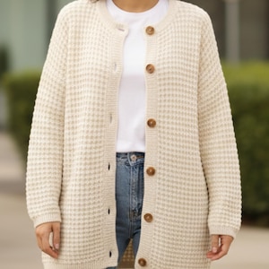 May include: A cream-coloured, long-sleeved cardigan with a waffle knit texture. The cardigan has a round neckline and is fastened with six brown buttons. The person is wearing a white t-shirt and blue jeans.