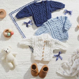 May include: An assortment of baby items, including three cable-knit sweaters in blue and white, a white teddy bear, brown leather baby shoes, and wooden toys. The sweaters have button closures and are arranged with blue ribbon bows.