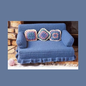 May include: A blue crocheted sofa with three square pillows. The sofa has rolled arms and a ruffled skirt. The pillows are square with colorful designs. A ball of pink yarn sits in front of the sofa.