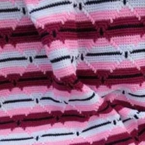 May include: A close-up of a crocheted blanket with a chevron pattern. The blanket features alternating rows of white, light pink, dark pink, and burgundy. Black lines separate the chevron rows. The blanket is folded, showing the texture.