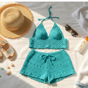 以下が含まれることがあります： ターコイズ色のクロシェ編みビキニセット。ホルターネックのトップスと紐付きのショートパンツが含まれ、麦わら帽子、サングラス、サンダル、日焼け止めと並べられています。白い表面に置かれたビーチウェアは、夏のテーマを連想させます。