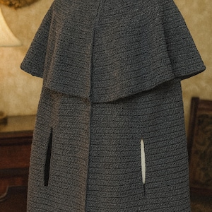 Op de afbeelding: Een grijze tweed cape met een gelaagd ontwerp. De cape heeft een korte, afgeronde kraag en twee armsplitten. De stof heeft een textuur en het kledingstuk is geschikt voor bovenkleding.