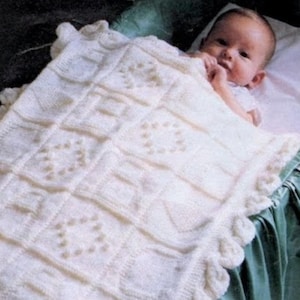 May include: A cream-colored, knitted baby blanket with a scalloped edge and a geometric pattern. The blanket is draped over a baby in a crib. The baby is lying on a white pillow.