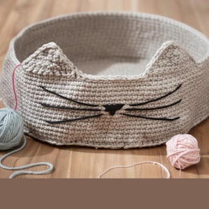 May include: A light gray, crocheted cat bed with cat ear details and black whiskers. The bed is round and sits on a wooden surface. Several balls of yarn in pink and blue are next to the bed, suggesting a handmade item.