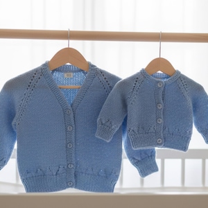 以下が含まれることがあります： 木製のハンガーに吊るされた、大人用と子供用の2つの水色のニットカーディガン。 カーディガンはボタン留めと装飾的なニットのディテールが特徴です。 背景は柔らかくニュートラルな色です。