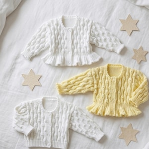 May include: Three baby cardigans in white and yellow, with a textured knit design and ruffled hems, are arranged on a white surface. Beige star-shaped decorations are scattered around the sweaters. The cardigans have long sleeves and button closures.