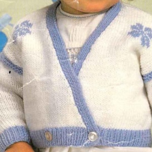 May include: A white and light blue knitted baby cardigan. The cardigan features a wrap-style design with light blue trim and leaf-like patterns on the shoulders. It has two small buttons for closure. The garment is made of a soft, textured yarn.