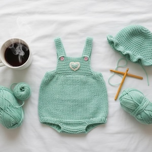 May include: A mint green knitted baby romper with a heart applique, a matching hat, and two balls of yarn. A cup of coffee and knitting needles are also present. The romper has pink buttons and is made of soft yarn.
