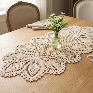 以下が含まれることがあります： クリーム色の、花の模様の入ったかぎ針編みのテーブルランナー。明るい色の木製のテーブルの上に置かれています。白い花が入った小さなガラスの花瓶が中央にあります。背景には2つの椅子が見えます。