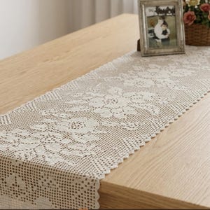 Op de afbeelding: Crèmekleurige, rechthoekige kanten tafelloper met een bloemmotief. De loper ligt op een lichtgekleurde houten tafel. Een fotolijst, een bloemenmand en een decoratieve klok zijn zichtbaar.