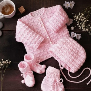 May include: A handmade, light pink crochet baby set, including a button-up cardigan, bonnet, and booties. The set is displayed with a cup of coffee, stones, and flowers on a dark wooden surface. The booties have a bow detail.