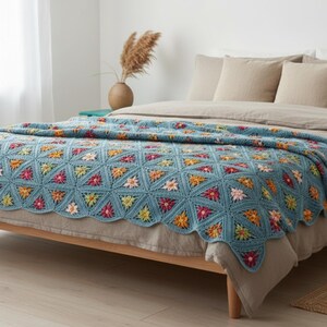 Könnte beinhalten: Eine blaue Häkeldecke mit Blumenmuster, die auf einem Bett liegt. Die Decke zeigt bunte Blumen in Rosa, Gelb und Orange. Das Bett hat einen hellen Holzrahmen und eine neutralfarbene Bettwäsche.