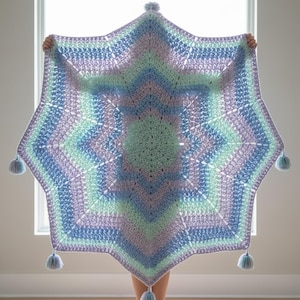 Peut inclure: Une grande couverture en crochet en forme d'étoile, aux couleurs pastel de bleu, lavande et vert menthe. La couverture présente un motif texturé et est ornée de pompons bleus clairs et doux à chaque pointe. La couverture est tenue devant une fenêtre.