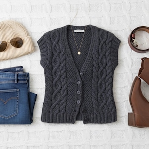 May include: An overhead shot of a casual outfit. It includes a gray cable-knit vest, blue jeans, brown ankle boots, a tan beanie, sunglasses, and a brown belt. A gold necklace with a pendant is also included.