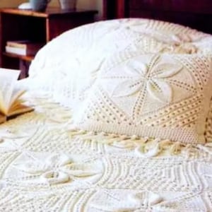 May include: A cream-colored, crocheted bedspread and pillow set. The pillow features a central floral design, surrounded by intricate openwork patterns. The bedspread and pillow are on a bed with a wooden headboard and a side table with a book.