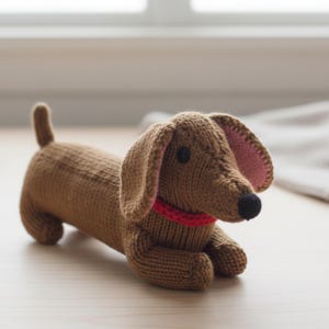 May include: A brown knitted dachshund dog toy with a red collar and black eyes. The dog has long floppy ears with pink inner ears and a small tail. The toy is lying down on a light-colored wooden surface.