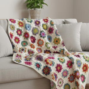 Puede incluir: Una manta de ganchillo hecha a mano con un patrón floral en varios colores, incluyendo rosa, amarillo, azul y verde. La manta está sobre un sofá gris con cojines a juego, creando un ambiente acogedor.