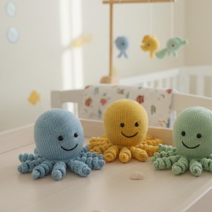 May include: Three knitted octopus toys in blue, yellow, and green sit on a light-colored surface. Each octopus has a round head, smiling face, and curled tentacles. The background includes a crib and a mobile with sea creature shapes.