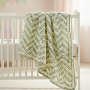 May include: A white crib with a soft mattress and a green and white chevron-patterned crochet blanket draped over the side. The blanket has a textured appearance and a decorative border. The crib is in a softly lit room.