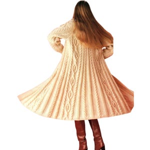 May include: A long, cream-colored cable-knit cardigan with a flared skirt. The sweater features intricate cable patterns and long sleeves. The person is wearing burgundy boots.